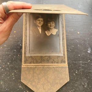 May include: Vintage black and white photograph of two children in a decorative frame. The frame is a light brown color with a textured pattern and a hinged cover. The photograph is in a rectangular shape.