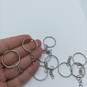 May include: A collection of silver-colored key rings with attached chains. The key rings are round and made of metal. The image is taken against a white background, highlighting the key rings. These are suitable for crafting or organizing keys.