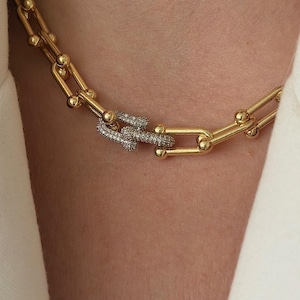Può includere: Una collana a catena color oro con maglie allungate a forma di U. Il centro della collana presenta una singola maglia incastonata con piccole pietre chiare e scintillanti, creando un punto focale distinto rispetto alle lisce maglie in metallo dorato.