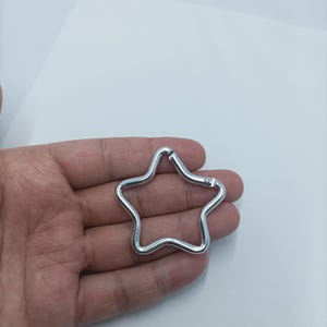 Puede incluir: Un llavero plateado en forma de estrella. El llavero es de metal y tiene un diseño sencillo. La estrella tiene cinco puntas y un cierre para sujetar llaves u otros objetos.