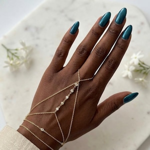 Könnte beinhalten: Ein goldenes Handkettchen mit einem filigranen Kettendesign, das ein Armband mit einer Fingerschlaufe verbindet. Der Schmuck ist mit mehreren kleinen, runden, klaren Steinen besetzt, die entlang der Mittelkette und am Handgelenk angeordnet sind und einen Lagen-Look erzeugen.