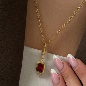 May include: A gold necklace featuring a delicate round link chain and a pendant with a rectangular red gemstone set in a gold frame. Below the red stone, a small triangular cluster of tiny clear stones hangs from the base of the pendant.