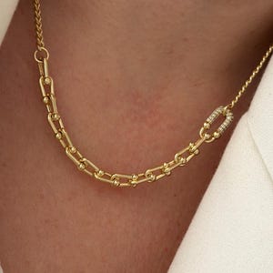 May include: A gold-toned chain necklace featuring elongated rectangular links with small spherical accents at each joint. One link is prominently encrusted with small, clear, sparkling stones, creating a distinct focal point on the chain.