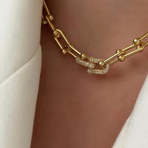 May include: A gold-toned chain necklace featuring elongated U-shaped links with small spherical accents at the joints. One central link is prominently encrusted with numerous tiny, clear, sparkling stones, creating a distinct focal point.