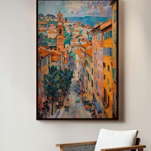 May include: An artistic print of a European cityscape, featuring a street lined with colorful buildings and a prominent church tower. The artwork uses warm tones, with orange rooftops and blue accents, creating a vibrant and inviting scene. The print is framed in a dark wooden frame.