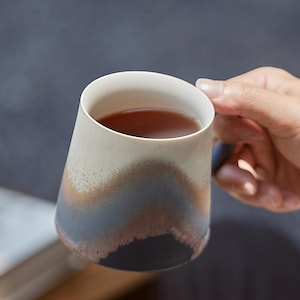 May include: A ceramic mug with a unique, layered design. The mug features a white rim, transitioning to shades of beige, brown, and blue. The mug is filled with a dark liquid, likely tea or coffee. The mug is held by a person.