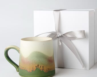 May include: A ceramic mug with a landscape design in shades of green, beige, and brown. The mug has a curved handle and is next to a white gift box tied with a silver ribbon. The mug is approximately 4 inches tall.