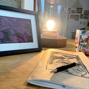 May include: A framed print of a comic book panel with purple and black ink. The panel features a character wearing a hat and the text "シエルダ" in Japanese. A notebook with a pencil and a floral mug are also in the image.