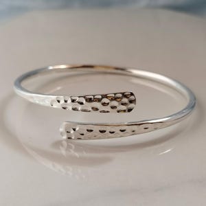 May include: A silver-colored cuff bracelet with a hammered texture on the ends. The bracelet has an open design, allowing it to be easily put on and taken off. The hammered texture adds a unique, handcrafted look to the jewelry.
