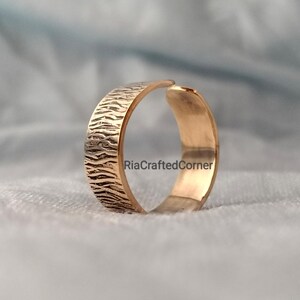 Puede incluir: Un anillo ajustable de color dorado con una banda texturizada. La banda presenta un patr&oacute;n detallado en forma de onda. El texto "RiaCraftedCorner" es visible en el anillo. El anillo est&aacute; sobre un fondo azul claro suave.