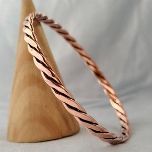 Könnte beinhalten: Ein kupferfarbenes Armreif-Armband mit einem gedrehten Seildesign. Das Armband wird auf einem kegelförmigen Holzobjekt präsentiert. Der Hintergrund ist hell und neutral. Das Armband ist ein schlichtes, elegantes Schmuckstück.