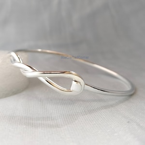 Puede incluir: Un brazalete plateado con un diseño de nudo. El brazalete está hecho de un metal brillante y reflectante, con un estilo simple y elegante. El nudo es el punto focal del brazalete.