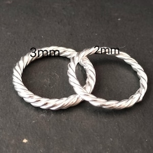 Op de afbeelding: Twee zilverkleurige, gedraaide metalen ringen. Eén ring is gemarkeerd met "3mm" en de andere met "2mm". De ringen zijn met elkaar verweven en tonen een gedetailleerde, touwachtige textuur. De achtergrond is donker en neutraal.