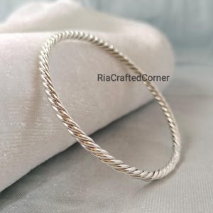 Può includere: Un bracciale rigido color argento caratterizzato da una texture a corda intrecciata continua lungo tutto il cerchio. La superficie metallica presenta una finitura lucida e riflettente. Il testo RiaCraftedCorner è visibile al centro.