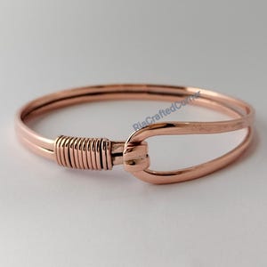 May include: A copper-coloured bracelet with a hook closure. The bracelet features a wrapped wire detail on one side and a curved, open design on the other. The metal has a polished finish.