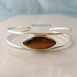 May include: A silver-toned cuff bracelet with a large, oval tiger's eye stone at the center. The stone is a warm brown color with natural striations. The bracelet has a minimalist design, with two parallel bands forming the cuff.