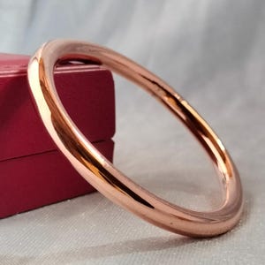 May include: A rose gold bangle bracelet. The smooth, polished bracelet has a circular design. The bracelet is displayed on a white surface with a red box in the background. The bangle is a simple, elegant piece of jewelry.