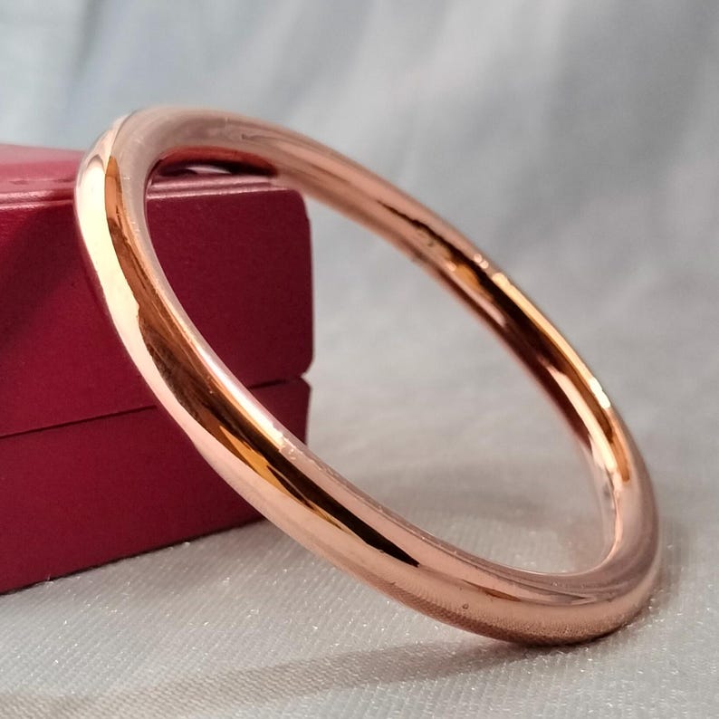 May include: A rose gold bangle bracelet. The smooth, polished bracelet has a circular design. The bracelet is displayed on a white surface with a red box in the background. The bangle is a simple, elegant piece of jewelry.