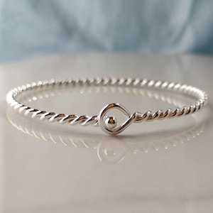 May include: A silver-colored twisted bangle bracelet. The bracelet features a delicate, rope-like design and a unique teardrop-shaped clasp with a small ball accent. The jewelry is displayed on a white surface with a soft blue background.