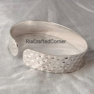 Puede incluir: Un brazalete de plata con textura martillada. El diseño abierto presenta una banda ancha con una serie de pequeñas hendiduras espaciadas uniformemente. El texto "RiaCraftedCorner" es visible.
