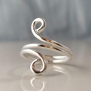 May include: A silver ring with a distinctive, swirling pattern. The ring has a continuous, flowing form that encircles the finger, giving it a stylish and contemporary appearance. The polished metal reflects light, accentuating the ring's complex curves.