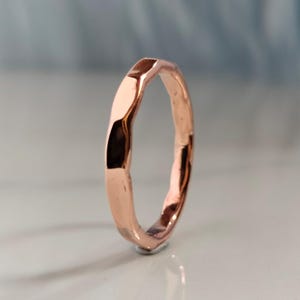 Puede incluir: Un anillo de oro rosa con un diseño facetado y texturizado. El anillo tiene un acabado pulido y una forma ligeramente irregular, que refleja la luz. El anillo está sobre un fondo suave y borroso.