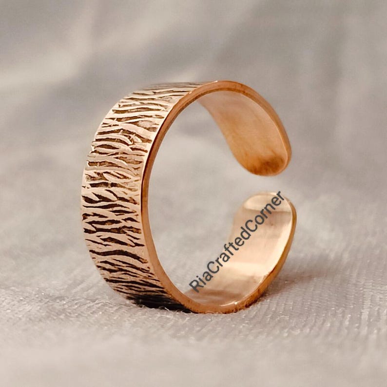 Puede incluir: Anillo abierto de oro rosa con una textura similar a la corteza. El dise&ntilde;o abierto permite el ajuste. El anillo se muestra sobre un fondo suave y neutro. El dise&ntilde;o del anillo es visible.