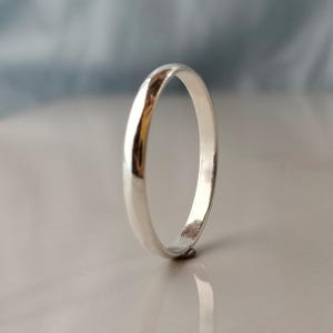 May include: A shiny, silver ring with a smooth, rounded band. The ring is displayed on a reflective surface, with a soft, blurred background. The ring's polished surface reflects light, creating highlights and shadows.