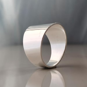 May include: A wide, polished silver ring with a smooth, curved design. The ring has a minimalist aesthetic and reflects light, creating a bright, shiny appearance. The band is thick and has a simple, elegant style.