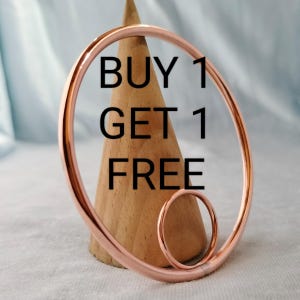 May include: Two copper-colored metal rings, one large and one small, are displayed in front of a wooden cone. The text "BUY 1 GET 1 FREE" is superimposed on the image in black letters. The rings have a polished, reflective surface.
