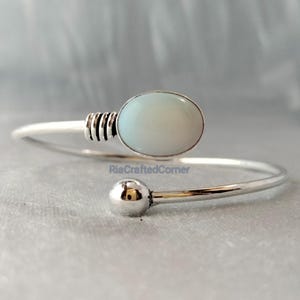 May include: A silver cuff bracelet featuring a pale blue oval gemstone. The bracelet has a minimalist design with a small ball on one end and a textured section near the gemstone. The text "RiaCraftedCorner" is visible.