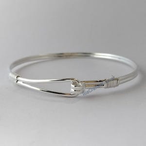 May include: A silver-colored bangle bracelet with a hook and eye closure. The bracelet is made of thin, round metal and has a simple, elegant design. The hook and eye closure is securely wrapped with wire. The bracelet is shown on a white background.