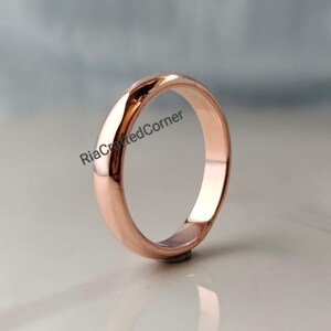 Peut inclure: Une bague anneau en or rose poli avec une surface lisse et arrondie. Le métal présente une teinte rose métallique chaude et une finition réfléchissante. Le texte "RiaCraftedCorner" est imprimé en lettres noires sur la partie supérieure de la bague.