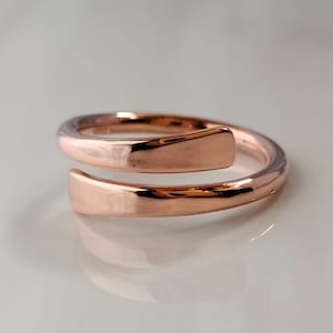 May include: A rose gold wrap ring with a smooth, polished finish. The ring has a spiral design with two flat, rectangular ends that overlap. The metal reflects light, creating highlights and shadows.