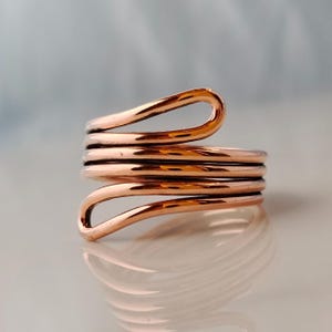 May include: A copper-coloured ring with a spiral design. The ring wraps around the finger multiple times, creating a layered effect. The metal has a polished finish, reflecting light. The ring's design is simple yet eye-catching, suitable for everyday wear.