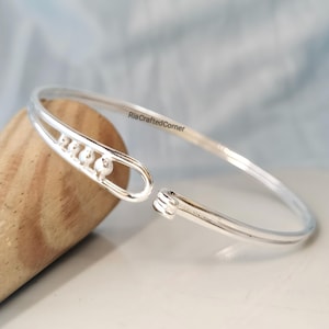 May include: A silver bangle bracelet with a distinctive design. The bracelet has a safety pin-style clasp and a decorative section with small, round embellishments. The text "RiaCraftedCorner" is visible on the bracelet.