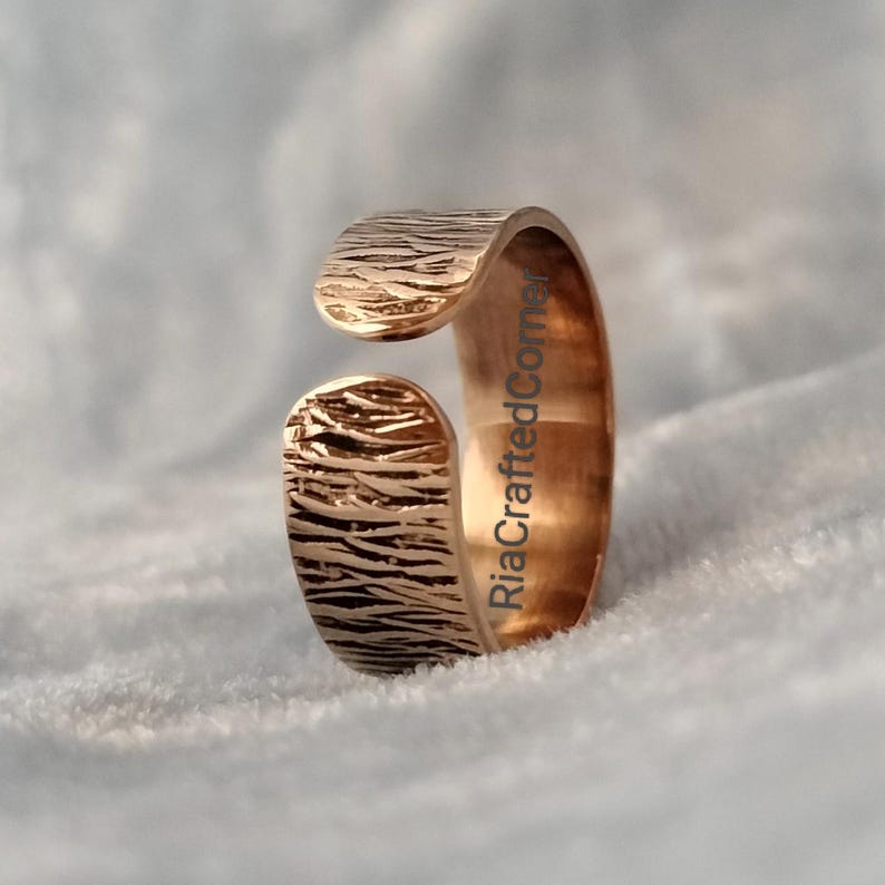 Puede incluir: Anillo abierto dorado con superficie texturizada. El anillo presenta un dise&ntilde;o org&aacute;nico con una textura similar a la corteza. El dise&ntilde;o abierto permite el ajuste de tama&ntilde;o. El texto "RiaCraftedCorner" es visible en el lateral del anillo.