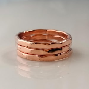Op de afbeelding: Drie koperkleurige ringen die op elkaar zijn gestapeld. Elke ring heeft een getextureerd, golvend oppervlak. De ringen worden gepresenteerd op een wit oppervlak, dat het licht reflecteert.