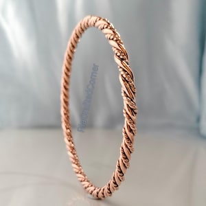 May include: A rose gold-colored bangle bracelet with a twisted rope design. The bracelet is circular and appears to be made of metal. The background is a soft, blurred blue and white.