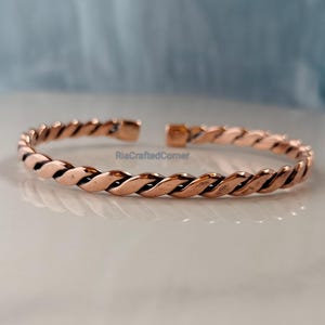 May include: A copper-colored, open cuff bracelet with a twisted rope design. The bracelet is made of a shiny metal and has a simple, elegant style. The bracelet is approximately 2.5 inches in diameter.