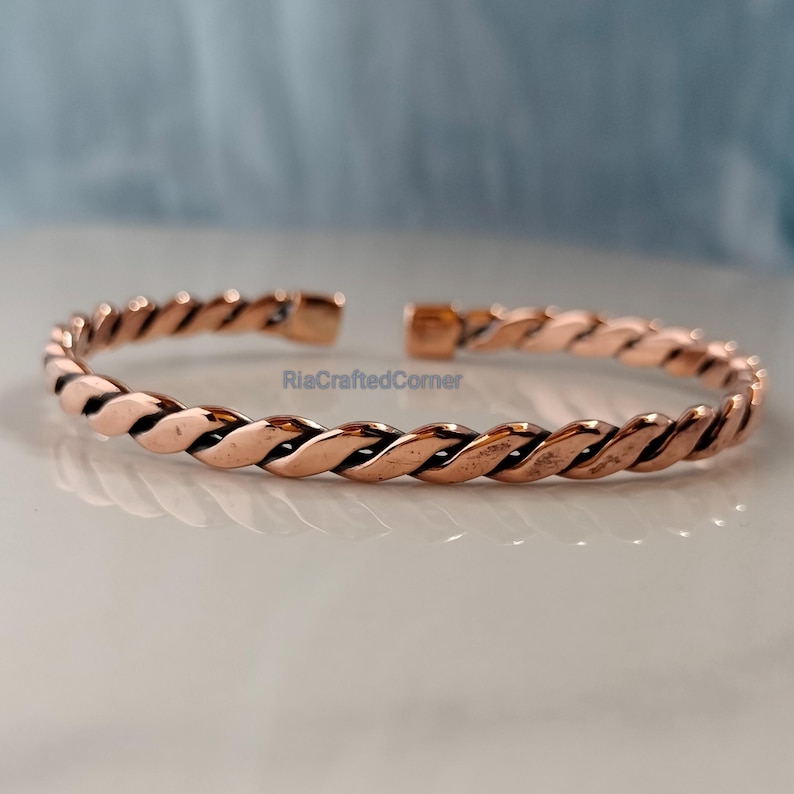May include: A copper-colored, open cuff bracelet with a twisted rope design. The bracelet is made of a shiny metal and has a simple, elegant style. The bracelet is approximately 2.5 inches in diameter.