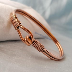 May include: A copper-colored bangle bracelet. The bracelet features a hook-and-eye closure and is accented with wrapped wire detailing. The metal has a polished finish, reflecting light. The bracelet is a simple, yet elegant piece of jewelry.