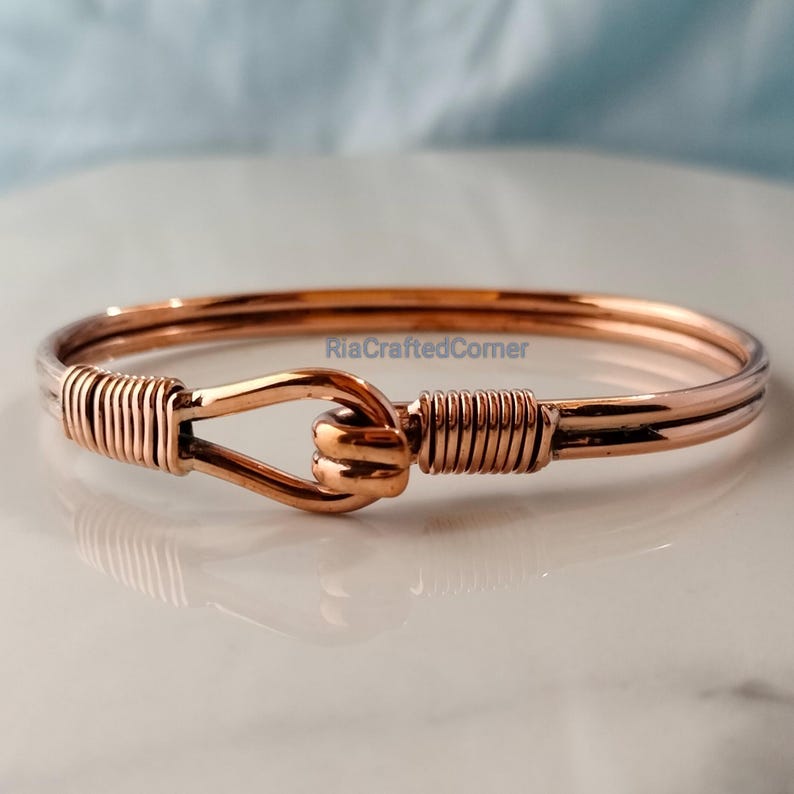 May include: A copper-colored bangle bracelet with a hook-and-eye clasp. The clasp is a stylized knot, and the ends of the bracelet are wrapped with copper wire. The bracelet is displayed on a white surface.