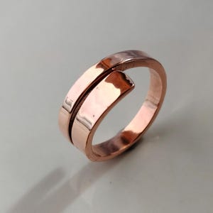Könnte beinhalten: Ein kupferfarbener Ring mit einem einzigartigen, offenen Design. Der Ring hat ein geteiltes Band, das sich umbiegt und einen offenen Raum schafft. Das Metall hat eine polierte, reflektierende Oberfläche.