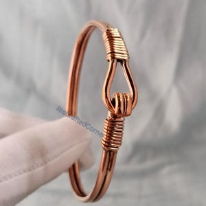 May include: A copper-colored bangle bracelet with a hook-and-eye closure. The bracelet features a wrapped wire design at the closure and on the lower portion of the hook. The bracelet is displayed on a white surface.