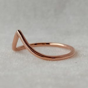 May include: A rose gold ring with a minimalist design. The ring has a smooth, curved band that rises to form a V-shape at the top. The ring is set against a soft, neutral background, highlighting its sleek, modern aesthetic.