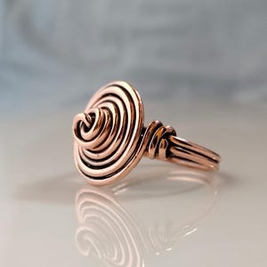 May include: A copper-colored ring with a spiral design. The ring features a circular, layered spiral at the center, with the band and connecting elements also made of copper wire. The ring is displayed on a reflective surface.