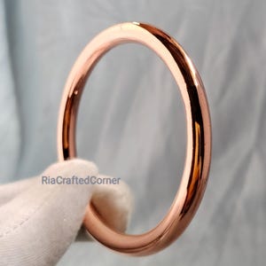 May include: A rose gold bangle bracelet. The smooth, circular bracelet reflects light, showcasing a polished finish. The bracelet is held by a gloved hand, with the text "RiaCraftedCorner" visible.
