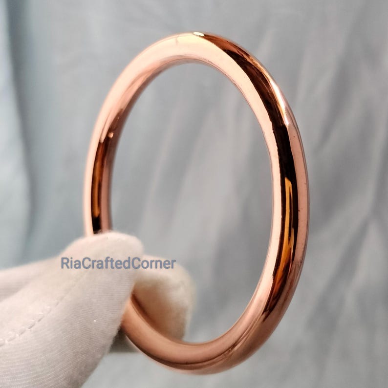May include: A rose gold bangle bracelet. The smooth, circular bracelet reflects light, showcasing a polished finish. The bracelet is held by a gloved hand, with the text "RiaCraftedCorner" visible.