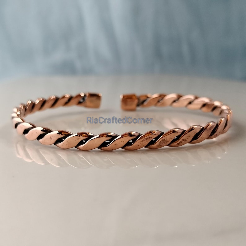 May include: A copper-colored, twisted-rope design cuff bracelet. The open-ended bracelet has a polished finish and is displayed on a white surface. The text "RiaCraftedCorner" is visible on the bracelet.